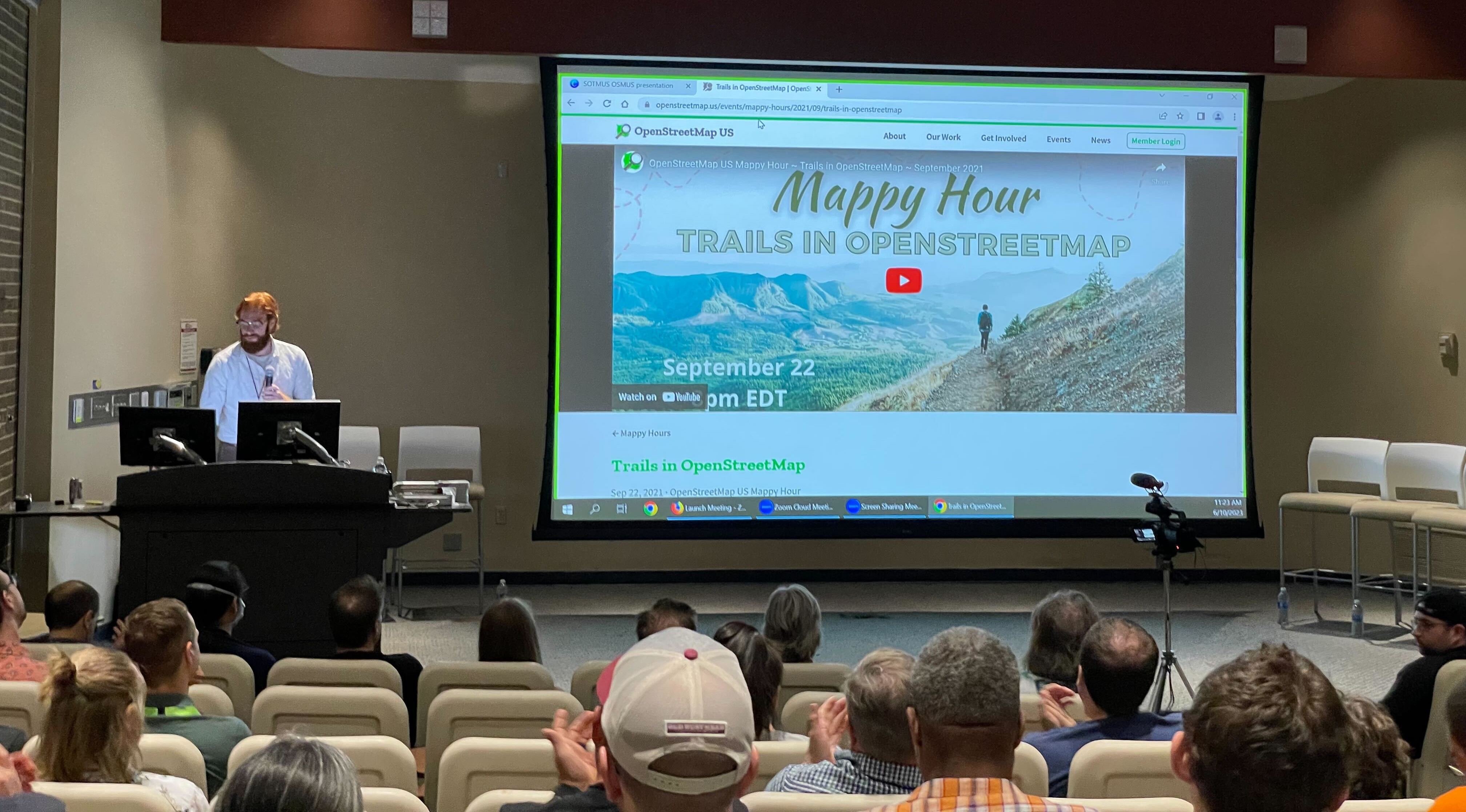 Quincy Morgan standing at a computer behind a podium in an auditorium. Onscreen is the OpenStreetMap US website open to the "Trails in OpenStreetMap" Mappy Hour page.